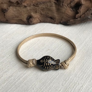May include: A simple bracelet made of beige cord with a black fish charm. The fish charm has gold detailing.