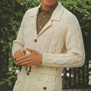 May include: A white cable knit cardigan with a brown belt and two pockets. The cardigan has a shawl collar and a button closure.