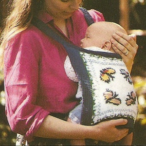 May include: A woman wearing a pink shirt is holding a baby in a white and blue knitted baby carrier with a butterfly design. The carrier has a blue and white striped strap.