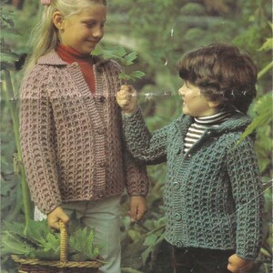 May include: Two children wearing handmade crochet jackets. One jacket is a dusty rose color, and the other is a teal green. The jackets have a button-up front and a textured, open weave design. One child holds a basket filled with leaves.