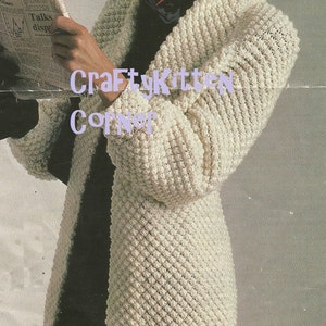 May include: Cream-colored, textured crochet coat with a wide collar. The coat has a diamond pattern and is knee-length. The image includes the text "CraftyKitten Corner" in purple. The person in the image is holding a newspaper.