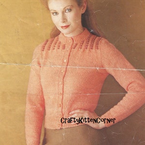 May include: A peach-coloured knitted cardigan with a button-up front and a beaded trim around the neckline. The cardigan is worn by a woman with red hair and a brown skirt.