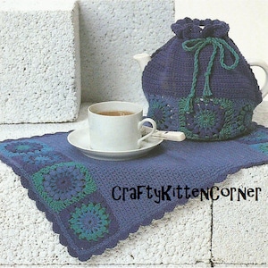 Puede incluir: Un calienta-tetera de ganchillo en azul y verde con un posavasos de ganchillo a juego y una taza de té blanca en la parte superior. El posavasos tiene un borde festoneado y el texto "CraftyKittenCorner" es visible en la esquina inferior derecha de la imagen.