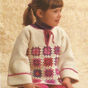 Puede incluir: Una niña pequeña lleva un suéter blanco de ganchillo con un patrón de cuadrados de la abuela rosa y morado. El suéter tiene un lazo en el cuello y ribetes rosas en las mangas. Lleva una falda rosa y tiene el pelo recogido en una coleta.