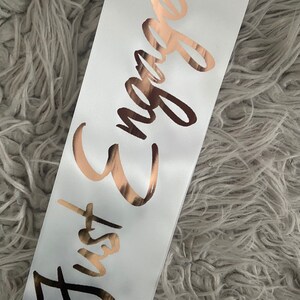 May include: White satin sash with the text "Just Engaged" in gold foil lettering.