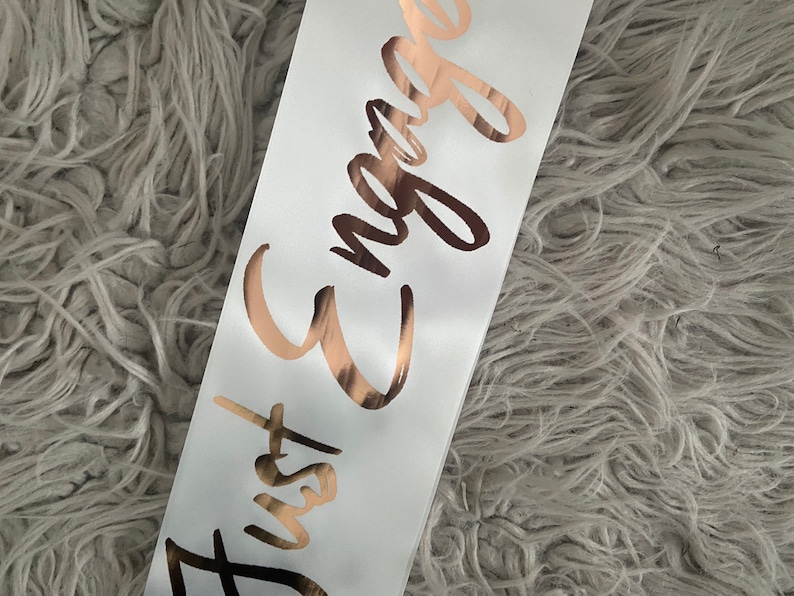 May include: White satin sash with the text "Just Engaged" in gold foil lettering.