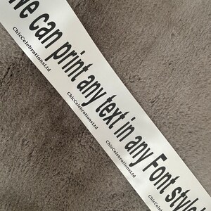 May include: White satin ribbon with the text "We can print any text in any Font style!" in black lettering. The text is repeated multiple times on the ribbon. ChicCelebrationsLtd is printed in smaller font below the text.