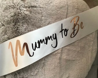 May include: White satin sash with the text "Mummy to Be" in black script. The "M" is gold foil.