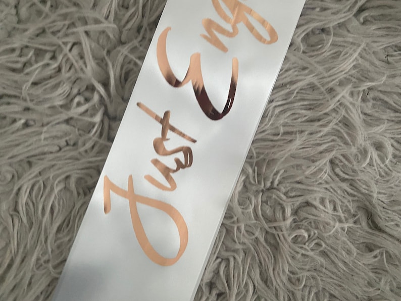 May include: White satin sash with the text "Just Engaged" written in gold foil lettering.