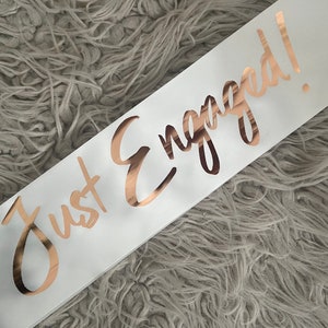 May include: White satin sash with the words "Just Engaged!" written in gold foil lettering.