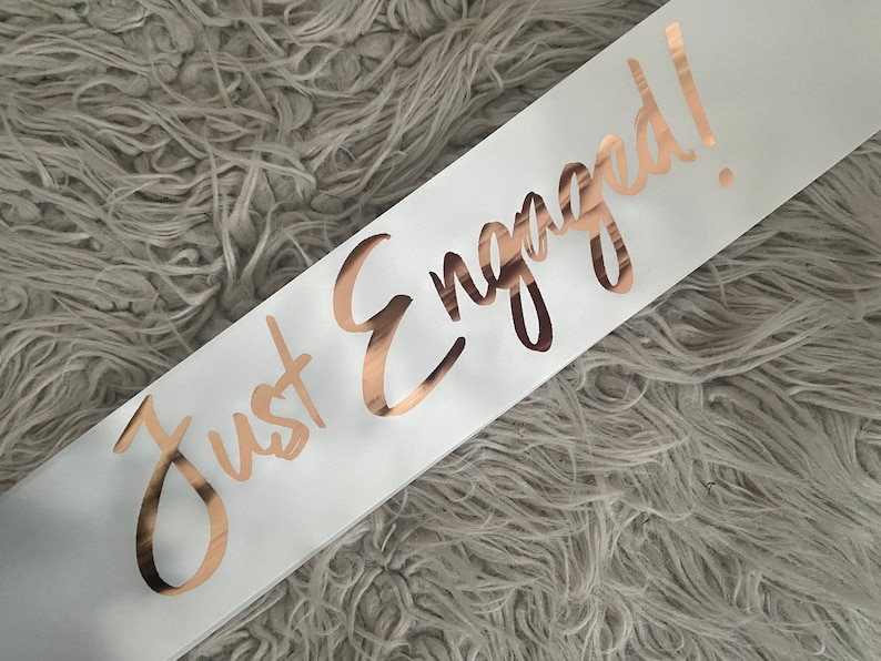 May include: White satin sash with the words "Just Engaged!" written in gold foil lettering.