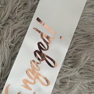 May include: White satin sash with the word "Engaged!" written in rose gold lettering.