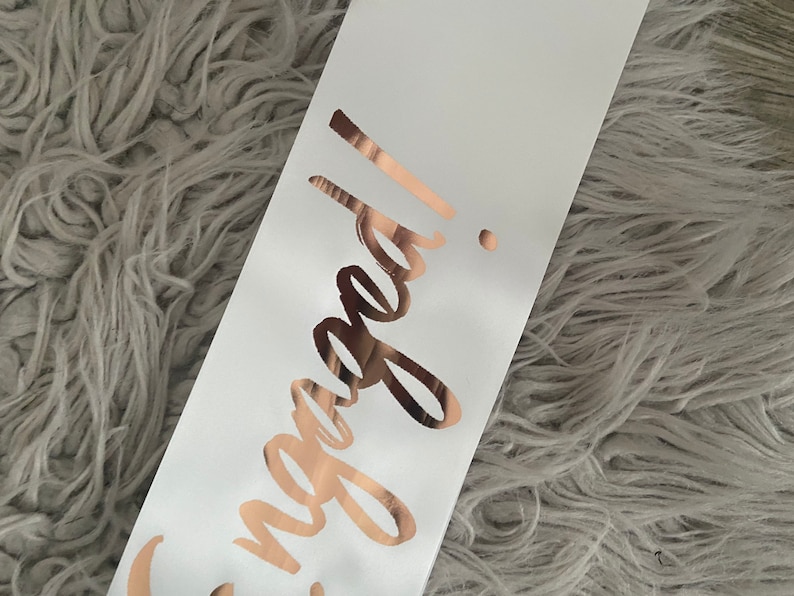 May include: White satin sash with the word "Engaged!" written in rose gold lettering.
