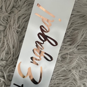 May include: White satin sash with the words "Engaged!" written in rose gold foil.