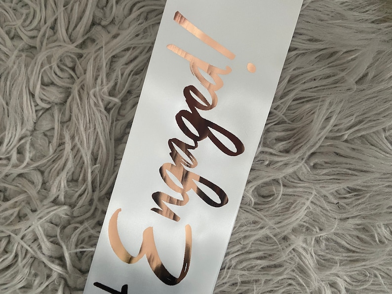 May include: White satin sash with the words "Engaged!" written in rose gold foil.