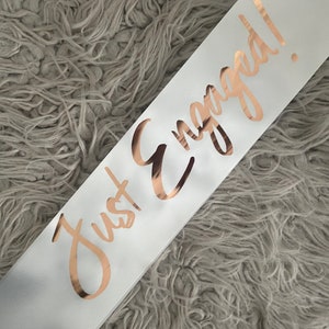 May include: White satin sash with the words "Just Engaged!" written in gold foil lettering.