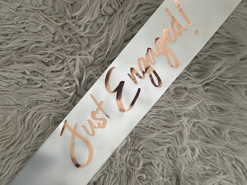 May include: White satin sash with the words "Just Engaged!" written in gold foil lettering.