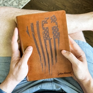 May include: A brown leather journal with a distressed American flag design and a cross. The journal has the name "John David" engraved on the front.