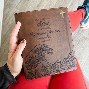 May include: A brown leather-bound journal with a gold cross on the cover. The journal features a black and white illustration of a wave and the text "HE STILLED THE storm TO A WHISPER, the waves of the sea WERE HUSHED Psalm 107:29" and "Carter James".