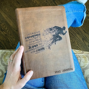 May include: A brown leather-bound journal with a black silhouette of a runner and the text "Let's run with endurance the race that is set before us, looking only to Jesus, the originator & perfecter of the faith Hebrews 12:1-2" and "Mike Johnson" on the cover.