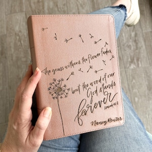 Puede incluir: Una cubierta de Biblia rosa con un diseño de diente de león y la cita "La hierba se seca, la flor se marchita, pero la palabra de nuestro Dios permanece para siempre" de Isaías 40:8. La cubierta se sostiene en el regazo de una persona.