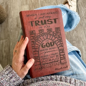 May include: A burgundy leather Bible cover with engraved text. The cover features the words "WHEN I AM AFRAID, I put my TRUST IN YOU" and "IN GOD" with a decorative archway design. Additional text includes a Bible verse and the name "Micah Hall."