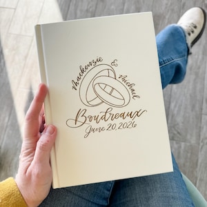 Pode incluir: Livro de visitas de casamento creme com nomes, anéis e data gravados. Os nomes "Mackenzie & Michael" e o sobrenome "Boudreaux" são exibidos acima da data "June 20, 2026". O livro está em uma mão.