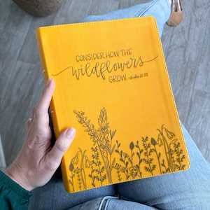 Puede incluir: Un diario amarillo con el texto "CONSIDER HOW THE WILDFLOWERS GROW. - Luke 12:27" y un dibujo de línea negra de flores silvestres en la parte inferior.