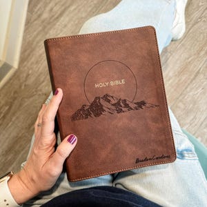 May include: A brown leather-bound Holy Bible with the words "HOLY BIBLE" above a mountain range design. The book is held in a person's lap. The name "Braden Carlons" is printed at the bottom right corner.