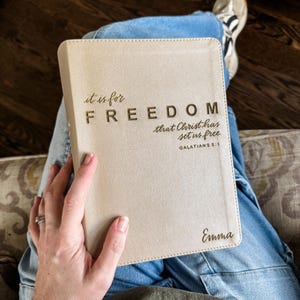 May include: A beige Bible cover with the words "it is for FREEDOM that Christ has set us free" and the name "Emma" printed on it. The text is in a dark brown color. The Bible is resting on a person's lap, with blue jeans and sneakers visible.
