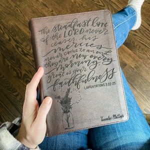 以下が含まれることがあります： 聖書の引用「主の揺るぎない愛は決して絶えることなく、その慈しみは尽きることがありません。それは毎朝新しく、あなたの忠実さは偉大です。」が刻まれた、茶色の革装丁のジャーナル。引用は筆記体で書かれており、ジャーナルは「Tessalei Plattner」という名前でパーソナライズされています。