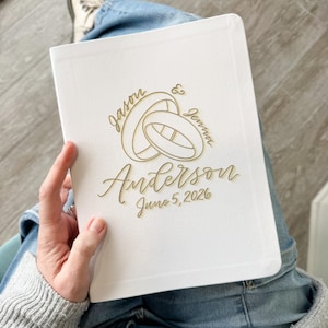 Puede incluir: Un libro de visitas de boda blanco con letras doradas. La portada presenta dos anillos entrelazados, los nombres "Jason" y "Jenna", el apellido "Anderson" y la fecha "5 de junio de 2026". El libro se sostiene en el regazo.