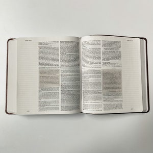 K&ouml;nnte beinhalten: Ein ge&ouml;ffnetes braunes Ledergebundenes Buch mit wei&szlig;en Seiten. Das Buch ist auf einer Seite mit dem Text "JOHN 19:18-19" und "JOHN 19:20-24" oben auf den Seiten ge&ouml;ffnet. Der Text ist in schwarzer Schrift und von einem wei&szlig;en Rahmen umgeben.