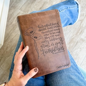Puede incluir: Una cubierta de Biblia de cuero marrón con el texto "El amor constante del Señor nunca cesa...grande es tu fidelidad" y una ilustración floral. La cubierta también incluye el nombre "Carla Fritzke".