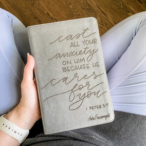 May include: A grey leather-bound journal with the words "cast all your anxiety on him because he cares for you" in cursive script. The text "I Peter 5:7" and "Ava Facemyer" are also printed on the journal.