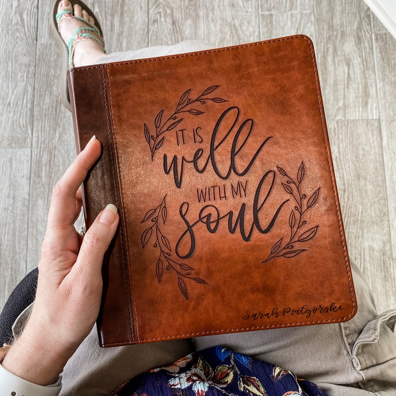 K&ouml;nnte beinhalten: Ein braunes, ledergebundenes Tagebuch mit den Worten "It is well with my soul" und einem floralen Design. Das Tagebuch ist mit dem Namen "Sarah Podgorski" personalisiert.