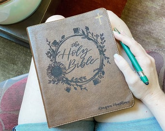 Puede incluir: Una Biblia de cuero marrón con las palabras "The Holy Bible" en escritura elegante, rodeada por una corona floral. Una cruz dorada está en la esquina superior derecha. El nombre "Reagan Matthews" está en la parte inferior. Un bolígrafo verde descansa sobre la Biblia.