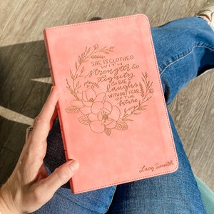 May include: A pink leather-bound journal with a gold floral design and the quote "She is clothed with strength & dignity, & she laughs without fear of the future." The journal is personalized with the name "Lucy Smith".