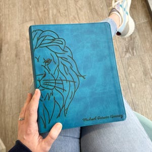 May include: A teal blue leather journal with a lion's face embossed on the front cover. The journal has the name "Michael Darwin Kenney" embossed on the bottom right corner.