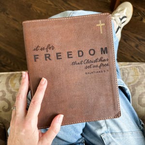 May include: A brown leather-bound book with the word "FREEDOM" embossed on the cover, along with the text "it is for" and "that Christ has set us free." A gold cross is in the upper right corner. The text "GALATIANS 5:1" is at the bottom.