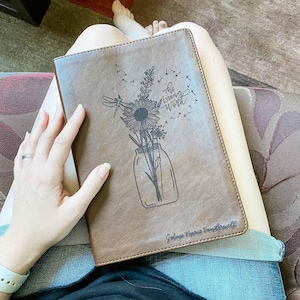 Peut inclure: Un journal relié en cuir marron avec un motif floral et les mots "The Living Word" gravés sur la couverture. Le journal est personnalisé avec le nom "Jolene Marie Vanderwell".