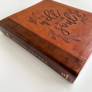 K&ouml;nnte beinhalten: Ein braunes, ledergebundenes Tagebuch mit goldenen Lettern, auf dem "Journaling Study Bible" steht. Der Einband zeigt ein gepr&auml;gtes Design eines Blattkr&auml;nzchens mit den Worten "It is well with my soul".