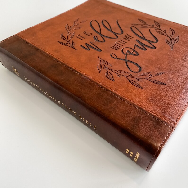 K&ouml;nnte beinhalten: Ein braunes, ledergebundenes Tagebuch mit goldenen Lettern, auf dem "Journaling Study Bible" steht. Der Einband zeigt ein gepr&auml;gtes Design eines Blattkr&auml;nzchens mit den Worten "It is well with my soul".