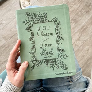 Op de afbeelding: Een saliegroen boek met de tekst "BE STILL & KNOW THAT I AM God" in zwart schrift, omgeven door bloemenlijntekeningen. Het boek is open, met de naam "Samantha Green" onderaan.