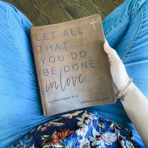 以下が含まれることがあります： 「あなたがたがするすべてのことを愛をもってしなさい」という言葉と聖書の引用「コリント人への手紙一 16:14」が書かれた茶色の革装丁のジャーナル。ジャーナルには、「ハンナ・ジュード・メリット」という名前も下部に印刷されています。