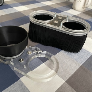 May include: A black brush attachment for a router, with a clear plastic base and two holes. The brush is attached to a clear plastic base with two screws. The brush is used for cleaning and sanding wood.