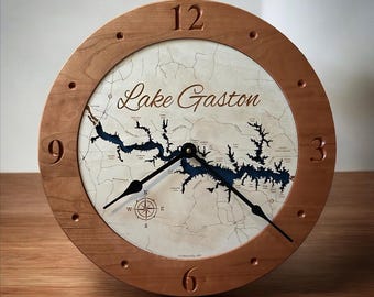 May include: A wooden clock with a map of Lake Gaston, North Carolina, on the face. The clock has a cherry wood frame and black hands. The numbers 3, 6, 9, and 12 are visible on the clock face.