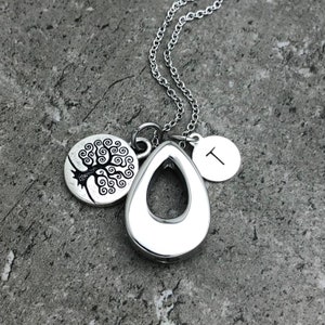 May include: Silver necklace with a teardrop-shaped pendant, a small round charm with a tree design, and a round charm with the letter "T" engraved on it.