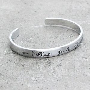 May include: Silver bangle bracelet with the inscription "follow your dreams" and an arrow pointing to the right.