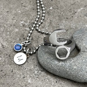May include: A silver-colored ring designed as a wrench, with a matching necklace featuring a blue gemstone and a circular pendant with the letter "M". The jewelry is displayed on a gray stone.